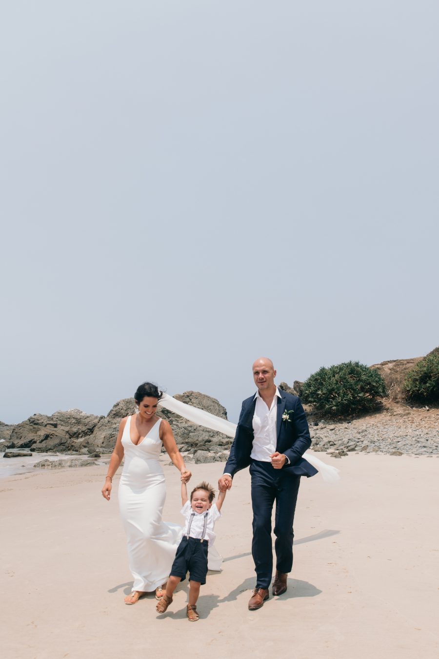 Bride and groom walk down beach with child / Broken Head Byron
