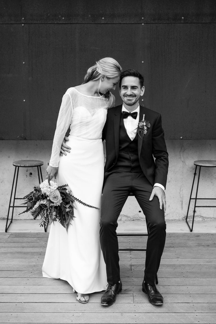 bride and groom smile with groom sitting and bride standing in stylish wedding at bay of fires bush retreat